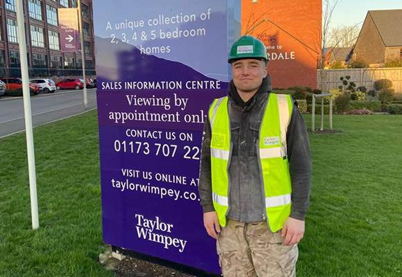 Keiran stood next to Taylor Wimpy development sign with high vis vest and helmet.